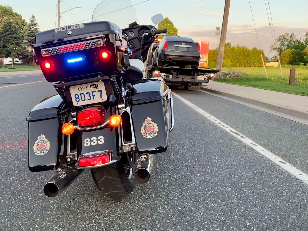 The rear-end of a Hamilton Police Service motorcycle on the side of the road with a vehicle on a flat-bed truck in the background.
